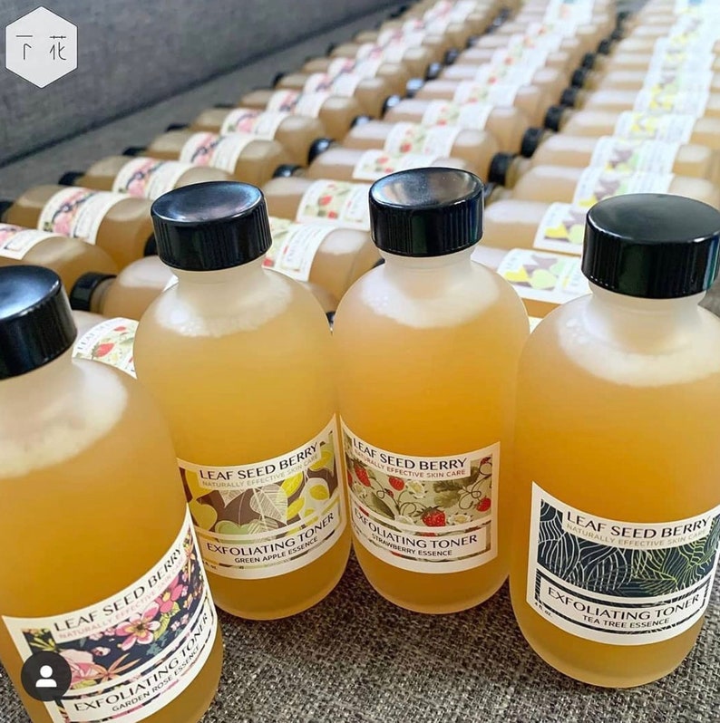 Pu&ograve; includere: Primo piano di diverse piccole bottiglie di vetro riempite con un liquido giallo chiaro. Le bottiglie hanno tappi neri ed etichette con il testo "Leaf Seed Berry" e "Exfoliating Toner" con diversi design a tema frutta.