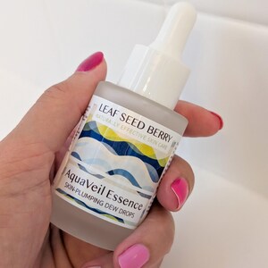 May include: A frosted glass bottle of AquaVeil Essence skin-plumping dew drops. The bottle has a white dropper top and a label with blue and yellow wave designs. The text "LEAF SEED BERRY" and "NATURALLY EFFECTIVE SKIN CARE" are also visible.