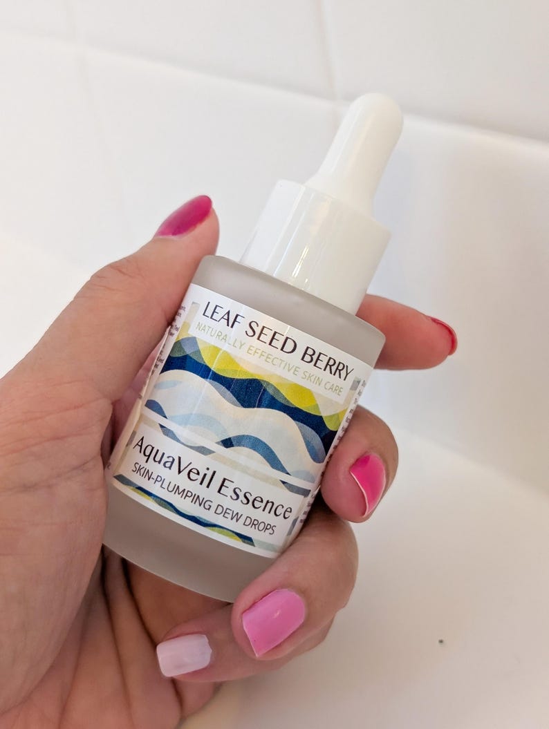 May include: A frosted glass bottle of AquaVeil Essence skin-plumping dew drops. The bottle has a white dropper top and a label with blue and yellow wave designs. The text "LEAF SEED BERRY" and "NATURALLY EFFECTIVE SKIN CARE" are also visible.