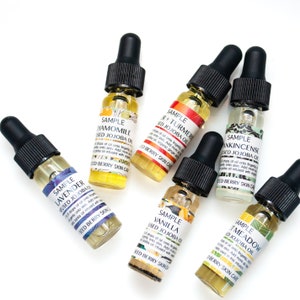 May include: Six small glass bottles with dropper tops filled with yellow liquid. Each bottle has a label with the words "Sample" and the name of a different essential oil blend: "Chamomile", "Lavender", "Vanilla", "Turmeric", "Meadow", and "Frankincense". The labels also include the words "Infused Jojoba Oil" and "Seed Berry Skin Care".