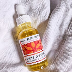 May include: A clear glass bottle of Leaf Seed Berry Ginger Turmeric infused jojoba oil face serum. The bottle has a white dropper and a label with orange floral design. The serum is yellow and the bottle is on a white towel.