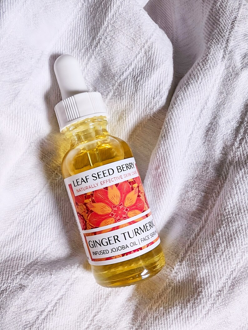 May include: A clear glass bottle of Leaf Seed Berry Ginger Turmeric infused jojoba oil face serum. The bottle has a white dropper and a label with orange floral design. The serum is yellow and the bottle is on a white towel.