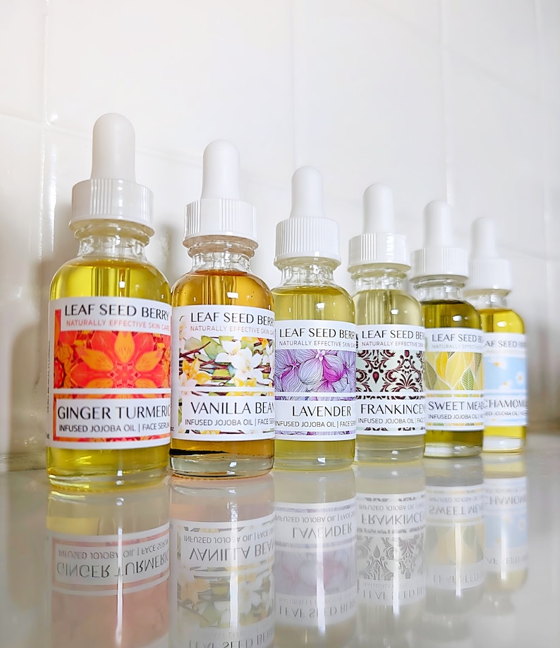 May include: A row of glass dropper bottles filled with yellow-tinted oil. Each bottle has a white dropper top and a label with botanical illustrations and text. The labels read "Leaf Seed Berry" and list various infused oils, including Ginger Turmeric and Vanilla Bean.