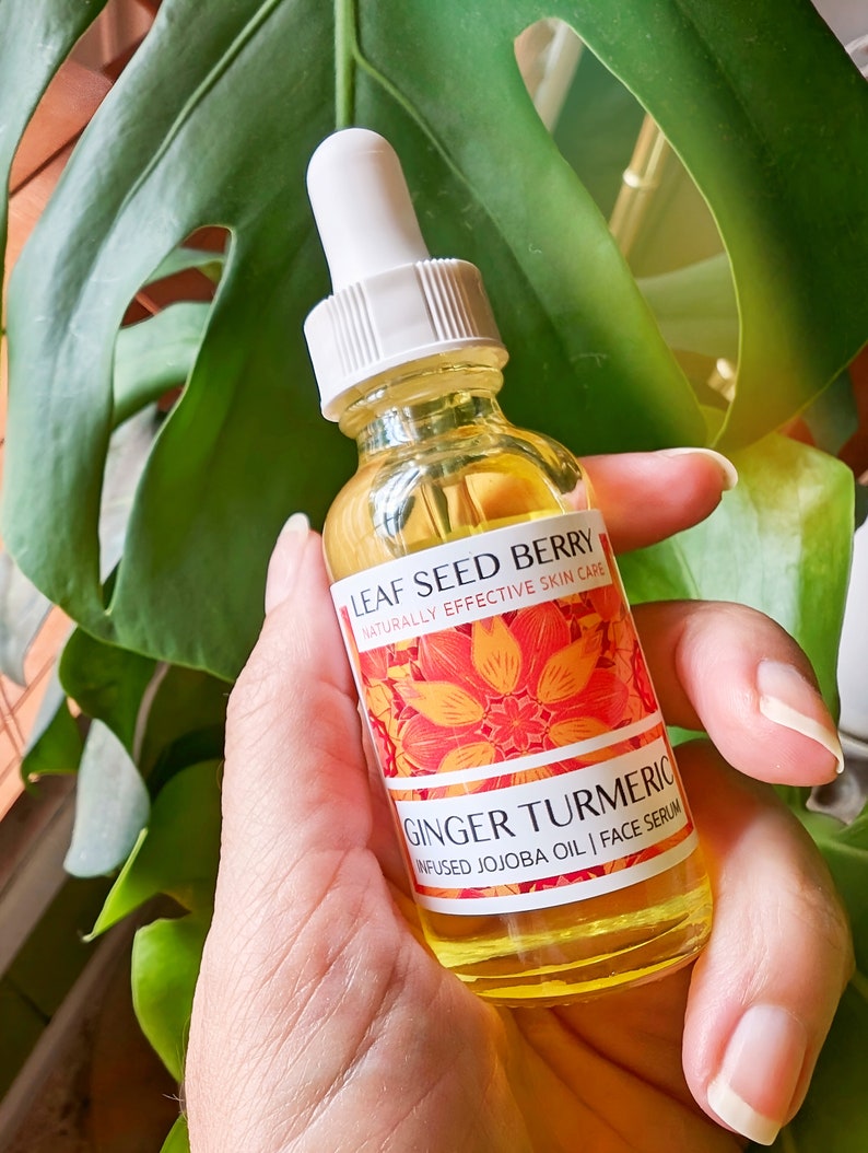 May include: A clear glass bottle of face serum with a white dropper is held in a hand. The label reads "Leaf Seed Berry" and "Ginger Turmeric Infused Jojoba Oil | Face Serum." The background features a large green leaf.