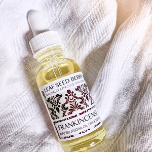 May include: A clear glass bottle with a white dropper top and a yellow liquid inside. The label is white with black text that reads "LEAF SEED BERRY NATURALLY EFFECTIVE SKIN CARE FRANKINCENSE INFUSED JOJOBA OIL | FACE SERUM".