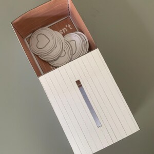 Puede incluir: Una caja de cart&oacute;n blanca con una ranura cortada en la parte delantera. La caja est&aacute; decorada con un patr&oacute;n de veta de madera. El interior de la caja es visible y contiene una pila de c&iacute;rculos de papel blanco con un coraz&oacute;n cortado en el centro. El texto "Isn't" es visible en el interior de la caja.