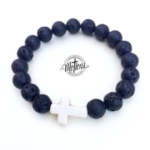 May include: A black lava stone bracelet with a white cross-shaped charm. The bracelet features dark, porous beads and a central white cross. The image includes the text "Motionz" in a circular design.