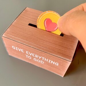 Puede incluir: Una caja de madera con una ranura en la parte superior. La caja es marr&oacute;n y tiene las palabras "Dar todo a Dios" impresas en la parte delantera. Una mano est&aacute; colocando una moneda amarilla con un coraz&oacute;n rojo en la ranura.