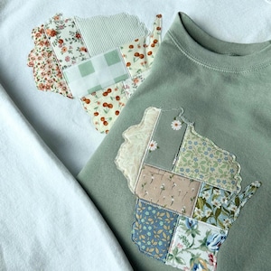 May include: Two sweatshirts, one white and one sage green, each featuring a patchwork design in the shape of the state of Wisconsin. The patchwork incorporates floral and patterned fabrics in various colors, including red, green, and blue. The sweatshirts are casual wear.