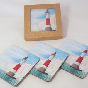Peut inclure: Un ensemble de quatre dessous de verre carrés représentant un phare à rayures rouges et blanches, une base marron et un sommet rouge. Le phare est placé devant des falaises blanches, de l'eau bleue et un ciel bleu clair. Un dessous de verre assorti est dans une boîte marron.