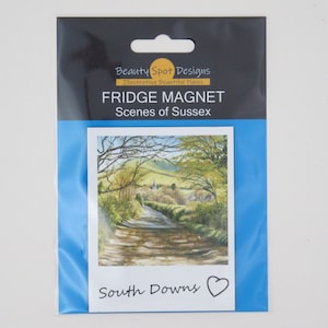 Peut inclure: Un aimant de réfrigérateur bleu et blanc avec une peinture à l'aquarelle d'une route sinueuse menant à un village dans les South Downs. L'aimant porte le texte "Beauty Spot Designs Illustrating Beautiful Places" et "Fridge Magnet Scenes of Sussex" en haut et "South Downs" en bas.