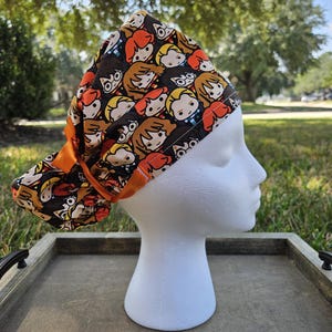 May include: A patterned scrub cap featuring cartoon character faces in shades of brown, orange, and black. The cap is on a white mannequin head, with an orange band and bow. The background includes green grass and trees.
