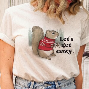 May include: A white t-shirt with a cartoon squirrel wearing a red sweater and holding a mug. The text "Let's get cozy." is printed on the shirt.