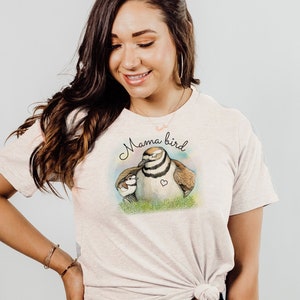 May include: A woman wearing a light beige t-shirt with a graphic of a bird and her chicks. The shirt says "Mama bird" in black text. She is wearing blue jeans.