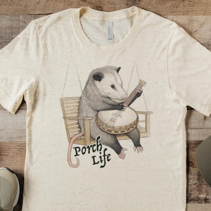 May include: Cream-colored t-shirt featuring a possum playing a banjo while seated on a wooden swing. The text "Porch Life" is below the possum. A green baseball cap and beige sneakers are also in the image.