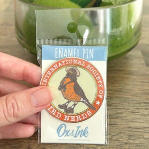 Puede incluir: Un pin de esmalte redondo que muestra un pájaro con sombrero, binoculares y zapatos, centrado en una tarjeta de respaldo con la etiqueta ENAMEL PIN INTERNATIONAL SOCIETY OF BIRD NERDS y Ox & Ink. El pin tiene un fondo crema con detalles en naranja y negro.