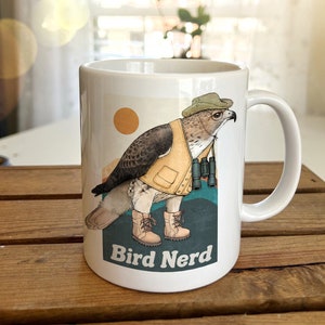 May include: White ceramic mug featuring a cartoon illustration of a hawk wearing a hat and vest, holding binoculars. The text "Bird Nerd" is printed on the mug.