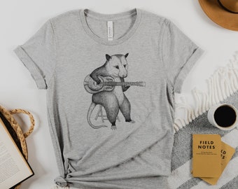 Könnte beinhalten: Ein graues T-Shirt mit einer Schwarzweißillustration eines Opossums, das Gitarre spielt. Das Opossum sitzt auf einem Hocker und hat einen überraschten Gesichtsausdruck.