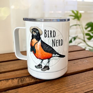 May include: White stainless steel travel mug featuring a black and orange bird wearing a hat and binoculars. The mug has the text "Bird Nerd" on it.