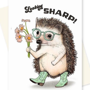 Peut inclure: Une carte de vœux illustrant un hérisson de dessin animé portant des lunettes vertes et des bottes de cow-boy, tenant un bouquet de fleurs roses et blanches. Le texte "Looking SHARP!" est écrit en noir sur la carte.