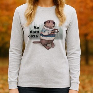 May include: A light gray long-sleeve shirt featuring a beaver wearing a sweater and holding a mug. The text "So dam cozy" is printed above the beaver. The background is a blurred image of fall foliage.