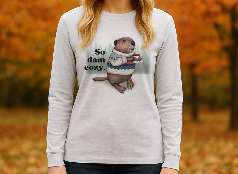 May include: A light gray long-sleeve shirt featuring a beaver wearing a sweater and holding a mug. The text "So dam cozy" is printed above the beaver. The background is a blurred image of fall foliage.