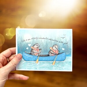 May include: A watercolor illustration of two otters paddling a canoe on a lake. The otters are wearing life jackets and holding paddles. The text "Life is an otter-ly wonderful adventure with you!" is written above the canoe.