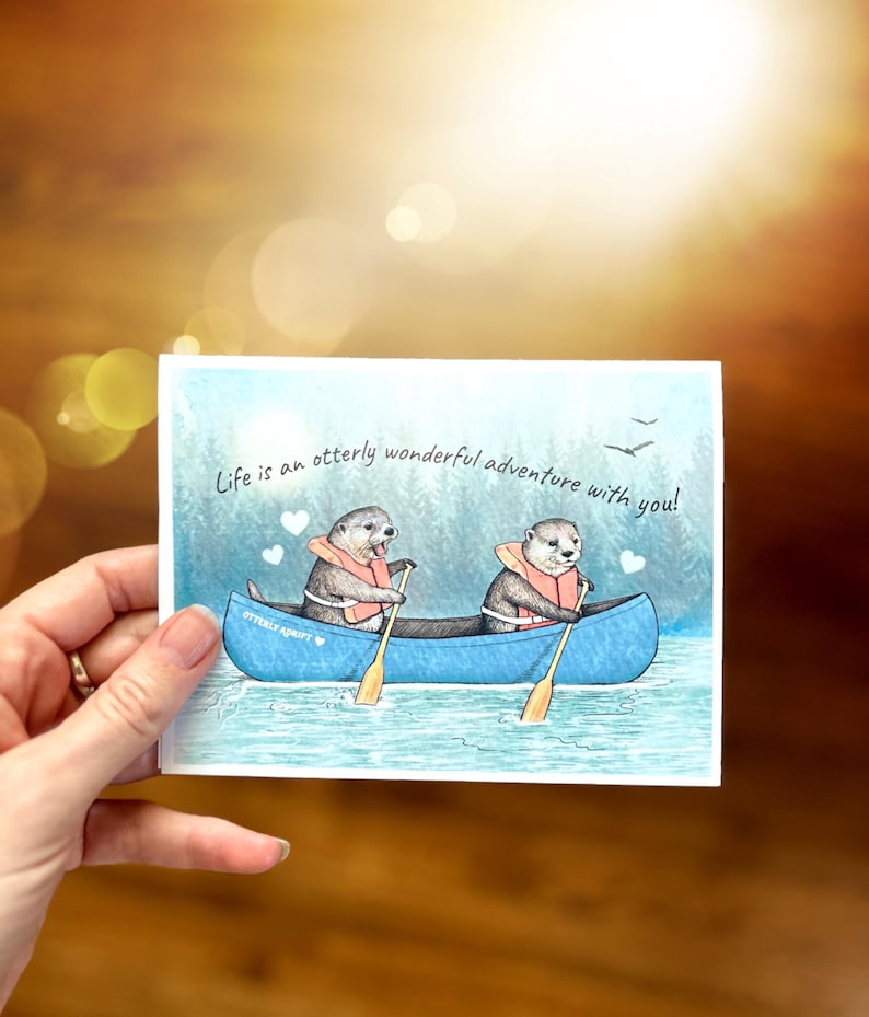 May include: A watercolor illustration of two otters paddling a canoe on a lake. The otters are wearing life jackets and holding paddles. The text "Life is an otter-ly wonderful adventure with you!" is written above the canoe.