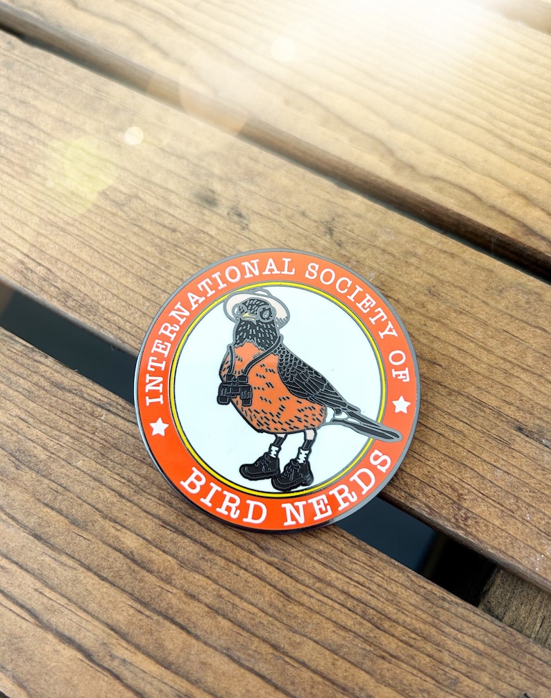 May include: A circular pin badge with an orange border and the text "INTERNATIONAL SOCIETY OF BIRD NERDS". The badge depicts a cartoon robin wearing a hat, binoculars, and boots. The bird is orange, black, and white.