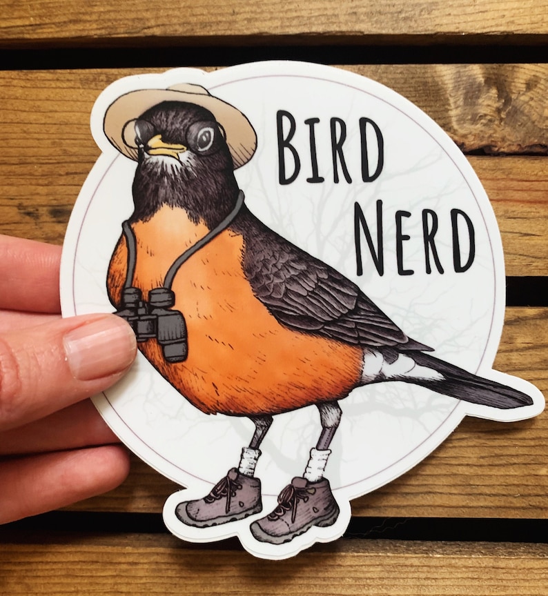 May include: A round sticker with a cartoon robin wearing a hat and binoculars. The robin is wearing hiking boots and has the text "BIRD NERD" on the sticker.