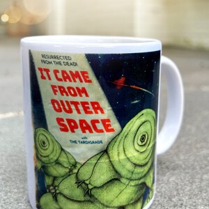 May include: White ceramic mug with a colorful graphic of a vintage sci-fi movie poster. The poster features the text "It Came From Outer Space" with the tagline "Resurrected From The Dead!" and "with The Tardigrade". The poster features a green alien creature with a large eye.