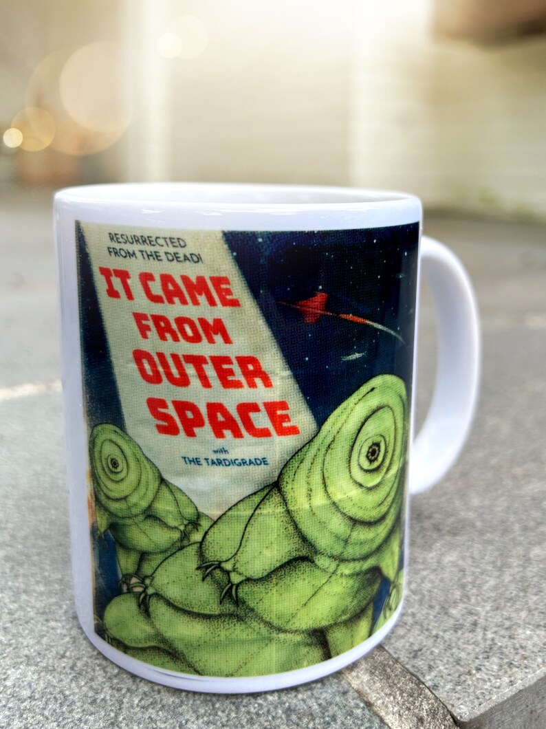 May include: White ceramic mug with a colorful graphic of a vintage sci-fi movie poster. The poster features the text "It Came From Outer Space" with the tagline "Resurrected From The Dead!" and "with The Tardigrade". The poster features a green alien creature with a large eye.