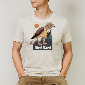 Può includere: Una maglietta bianca con una grafica di un uccello che indossa un cappello e tiene un binocolo. Il testo "Bird Nerd" è stampato sotto la grafica.