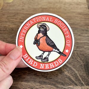 May include: A circular sticker featuring a robin wearing a tan hat, hiking boots, and binoculars around its neck. The bird is centred within a cream-coloured circle surrounded by a red border with the text INTERNATIONAL SOCIETY OF BIRD NERDS.