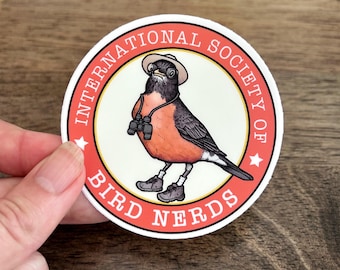 Puede incluir: Una pegatina circular que muestra un petirrojo con un sombrero beige, botas de senderismo y binoculares al cuello. El ave está centrada en un círculo color crema rodeado por un borde rojo con el texto INTERNATIONAL SOCIETY OF BIRD NERDS.
