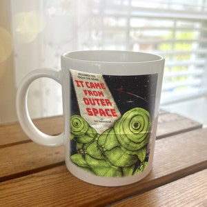 May include: White ceramic mug with a graphic of a vintage movie poster for "It Came From Outer Space" featuring a green alien creature. The poster text reads "Resurrected From The Dead" and "The Tardigrade".
