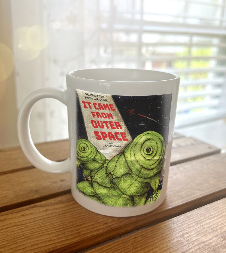 May include: White ceramic mug with a graphic of a vintage movie poster for "It Came From Outer Space" featuring a green alien creature. The poster text reads "Resurrected From The Dead" and "The Tardigrade".