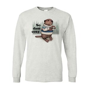 May include: A light gray long-sleeve shirt featuring a cartoon beaver wearing a sweater and holding a mug. The text "So dam cozy" is printed on the shirt. The beaver is brown and the sweater has blue and white accents.