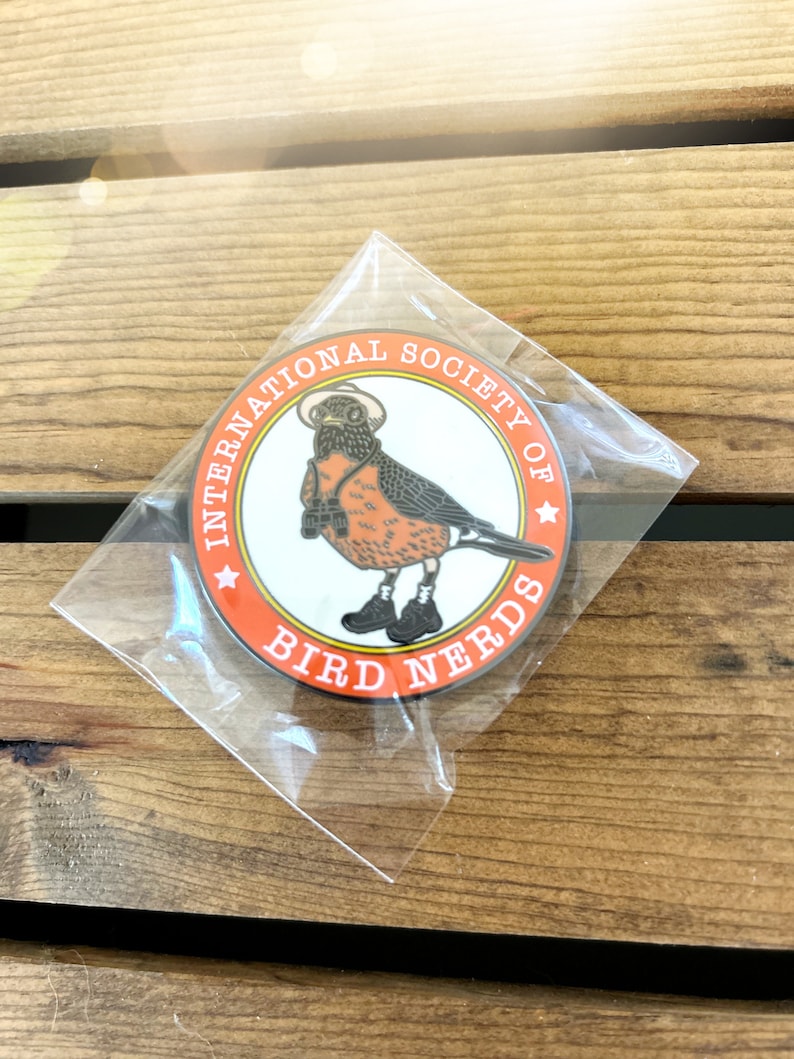 May include: A circular pin badge with a cartoon robin wearing a hat, glasses, and boots, holding binoculars. The badge is orange and white and reads "INTERNATIONAL SOCIETY OF BIRD NERDS". It is in a clear plastic packet.