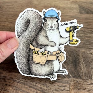 May include: A die-cut sticker featuring a detailed illustration of a squirrel wearing a light blue hard hat and a tan tool belt filled with various tools. The squirrel holds a yellow power drill and the text Ahhh nuts! is printed in black.
