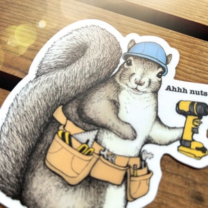 Peut inclure: Un &eacute;cureuil de dessin anim&eacute; portant un casque bleu et une ceinture &agrave; outils tient une perceuse jaune et noire. L'&eacute;cureuil regarde la perceuse et a une bulle de dialogue qui dit "Ahhh nuts".