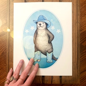K&ouml;nnte beinhalten: Eine Cartoon-Illustration eines Otters, der einen blauen Cowboyhut und blaue Stiefel tr&auml;gt und auf einem blauen und wei&szlig;en Hintergrund mit wei&szlig;en Sternen steht.