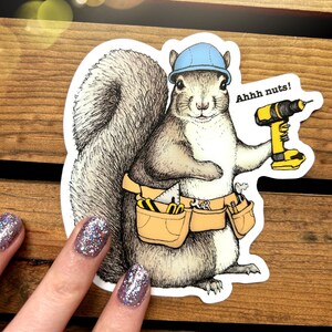 Peut inclure: Un &eacute;cureuil de dessin anim&eacute; portant un casque bleu et une ceinture &agrave; outils tient une perceuse jaune et noire. L'&eacute;cureuil regarde le spectateur et le texte "Ahhh nuts!" est au-dessus de la perceuse.