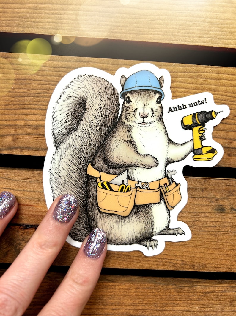 Peut inclure: Un &eacute;cureuil de dessin anim&eacute; portant un casque bleu et une ceinture &agrave; outils tient une perceuse jaune et noire. L'&eacute;cureuil regarde le spectateur et le texte "Ahhh nuts!" est au-dessus de la perceuse.