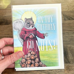 Puede incluir: Una tarjeta de felicitación con una ardilla vestida con una túnica roja y un halo amarillo detrás de la cabeza. La ardilla sostiene una bellota y hay un montón de bellotas a sus pies. El texto dice ON THY BIRTHDAY GO NUTS! sobre un cielo azul.