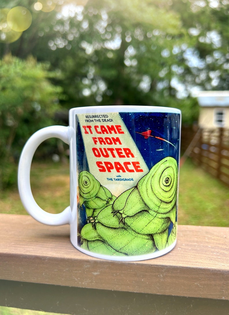 May include: White ceramic mug with a colorful graphic of a creature from outer space. The graphic is a vintage-style poster with the text "It Came From Outer Space with The Tardigrade".