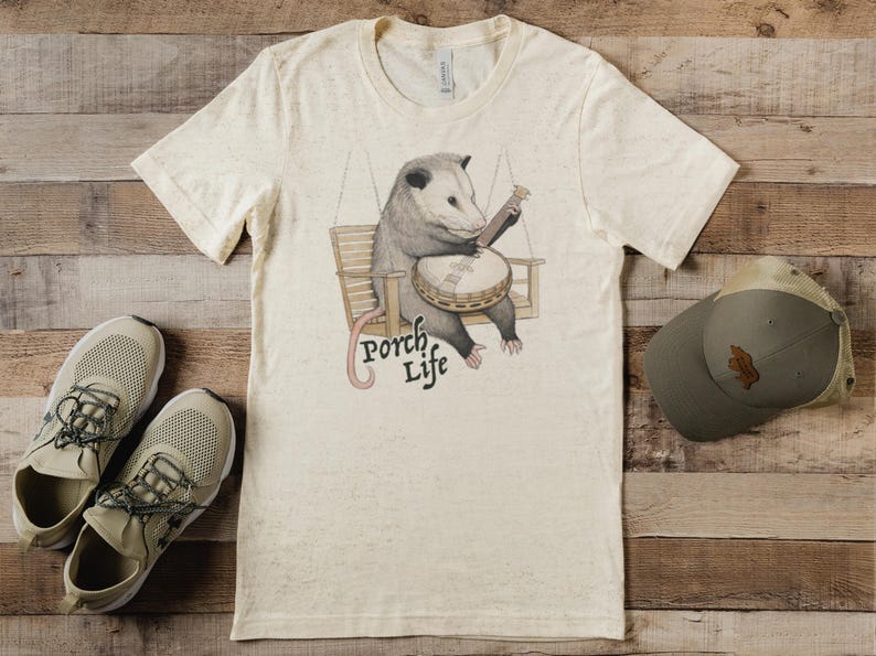 May include: Cream-colored t-shirt featuring a possum playing a banjo while seated on a wooden swing. The text "Porch Life" is below the possum. A green baseball cap and beige sneakers are also in the image.