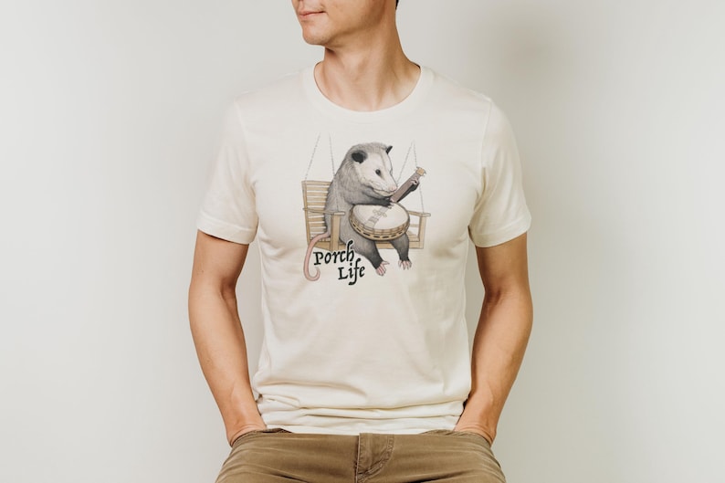 May include: Cream-colored t-shirt featuring a cartoon opossum playing a banjo while seated on a porch swing. The text "Porch Life" is printed below the illustration in a whimsical font. The shirt is made of a soft material.