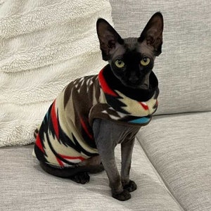 May include: A black hairless cat wearing a brown, red, white, and blue patterned fleece sweater. The cat is sitting on a light gray couch.