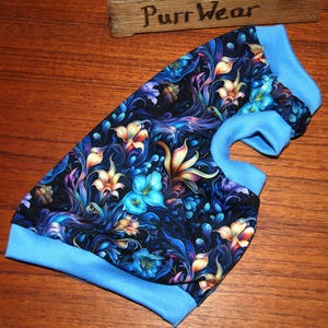 May include: A dog shirt with a vibrant floral pattern in shades of blue, purple, and orange. The shirt has light blue trim around the neck and sleeves. The wooden sign in the background reads "Purr Wear."